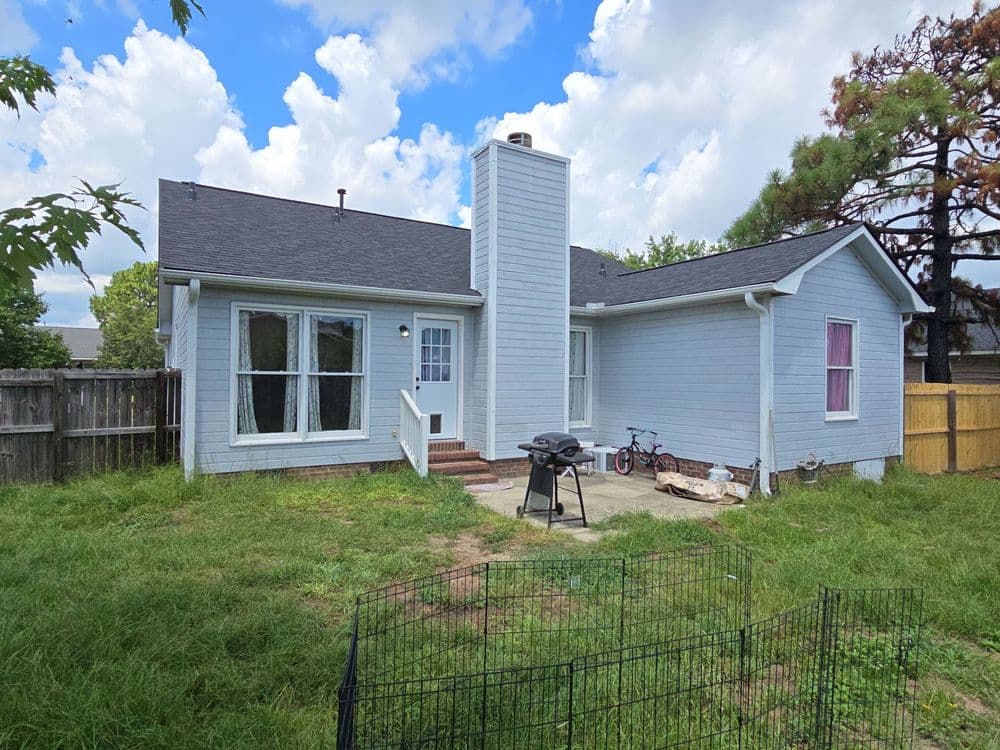 Single-story blue house with backyard, grill, and bike, surrounded by greenery and fence.