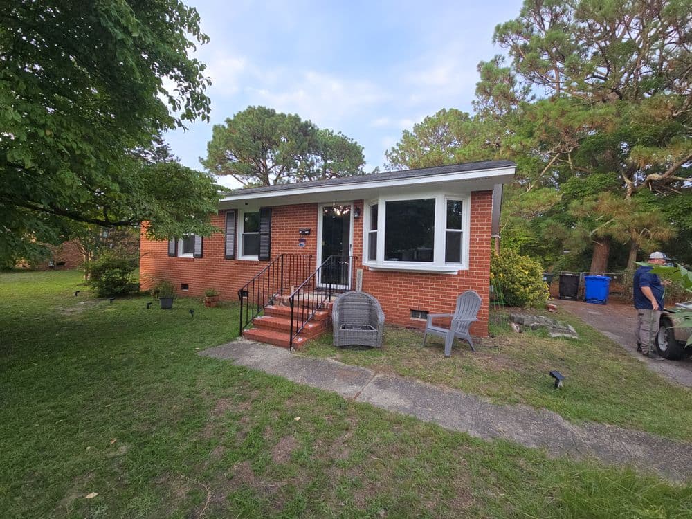 Brick house with steps, front yard, and patio chairs surrounded by trees and a driveway.