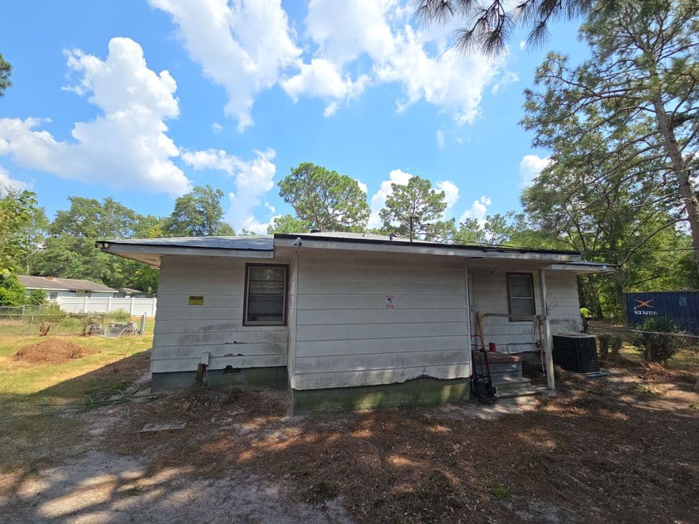 Dilapidated house in a wooded area under a partly cloudy sky with overgrown yard.