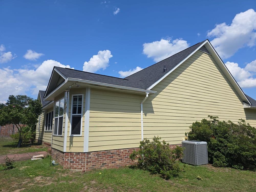Yellow house exterior with brick foundation, lush greenery, and clear blue sky in background.