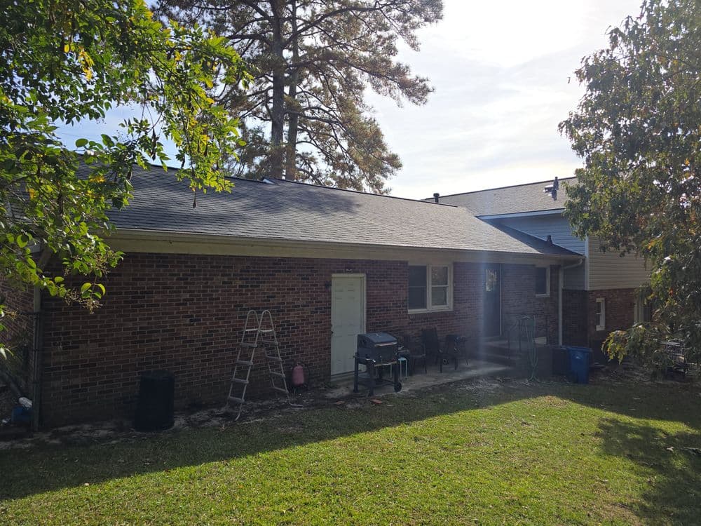 Brick home backyard with grill, ladder, trees, and green lawn under clear skies.