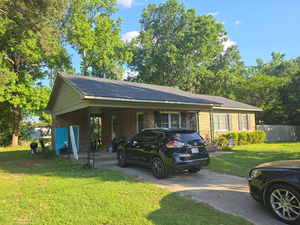 Brick house with a carport, black SUV parked on the driveway, surrounded by green trees.