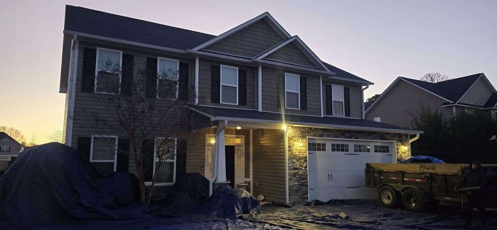 Modern two-story house with stone accents, illuminated porch, and blue tarps covering the yard.