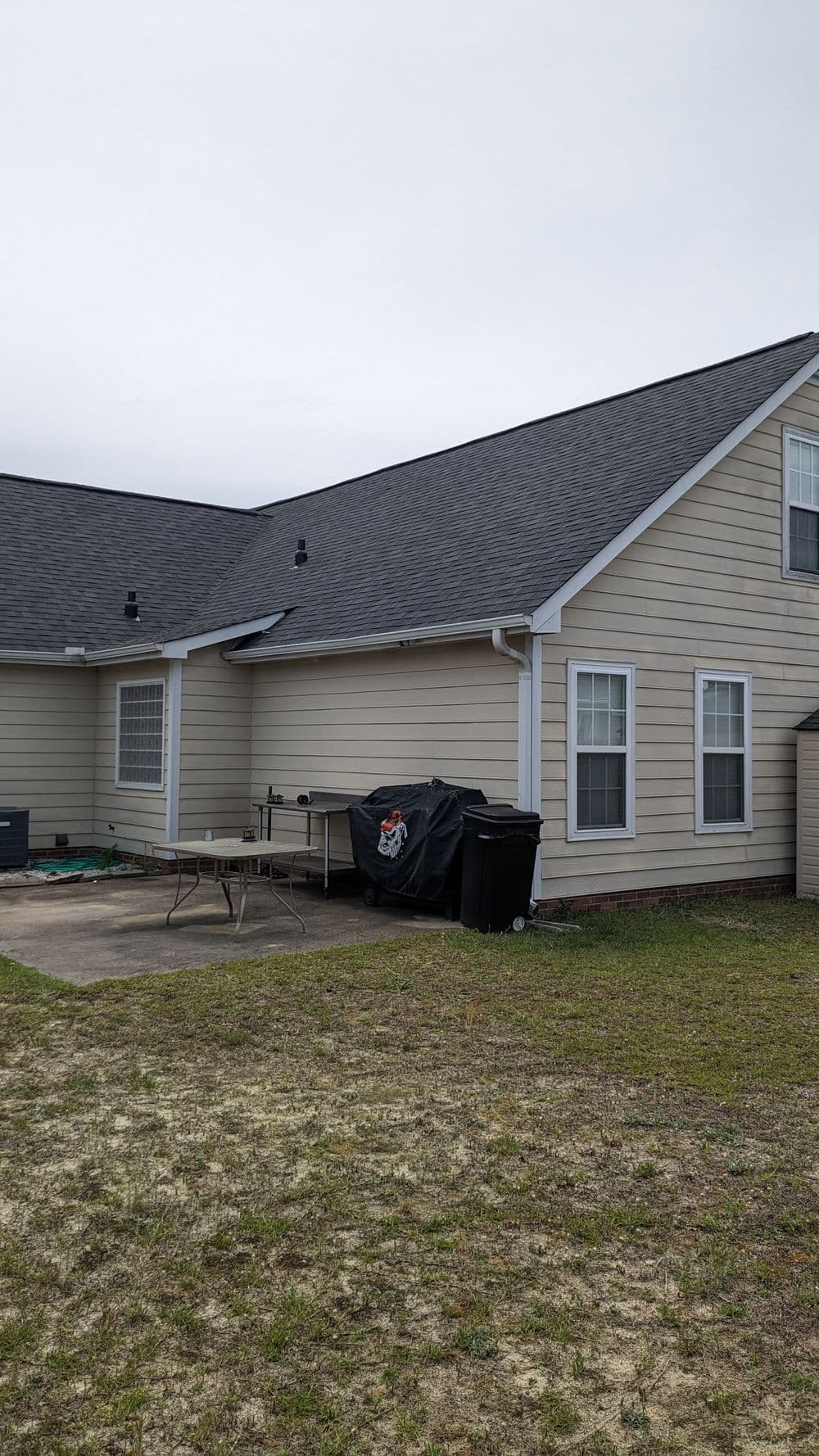 Back view of a house with a patio, table, grill cover, and trash bins on a grassy lawn.