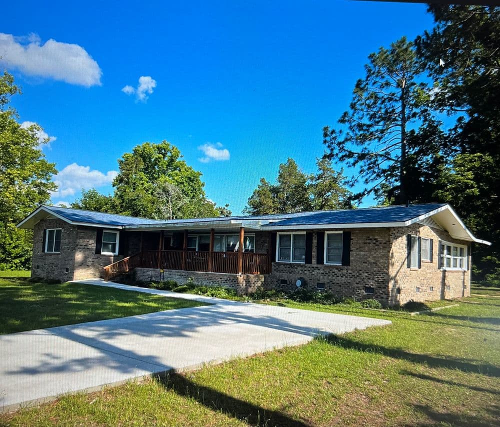 Single-story brick house with a porch, surrounded by green trees and blue sky.