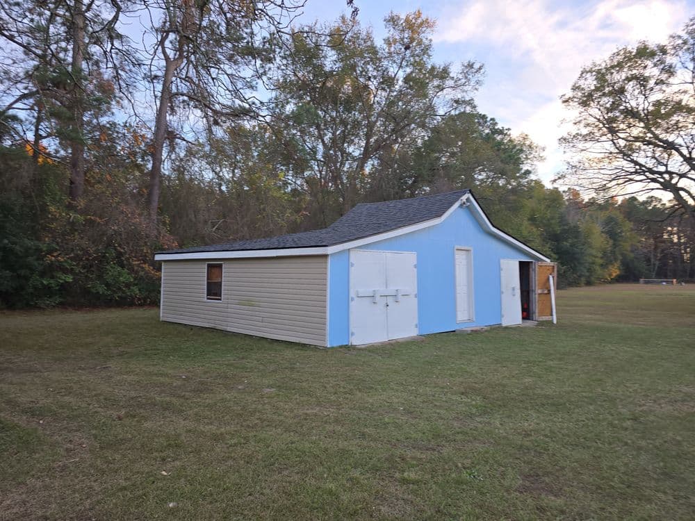 Blue-sided storage shed in a grassy field surrounded by trees under a clear sky.