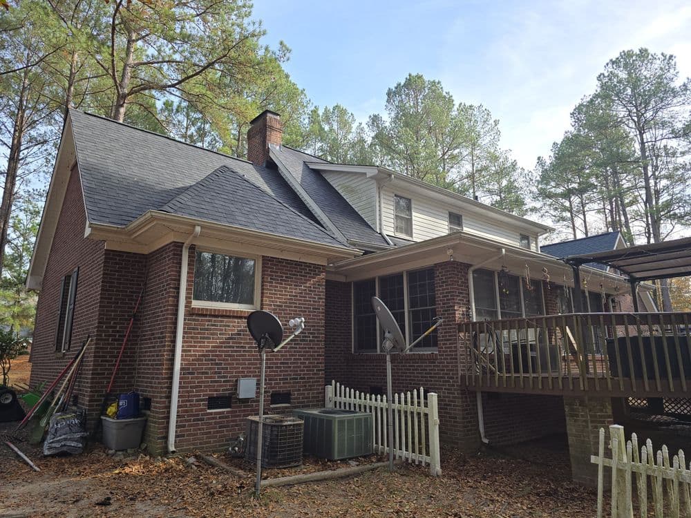 Brick house exterior with a deck, satellite dish, and wooded background. Rural residential setting.
